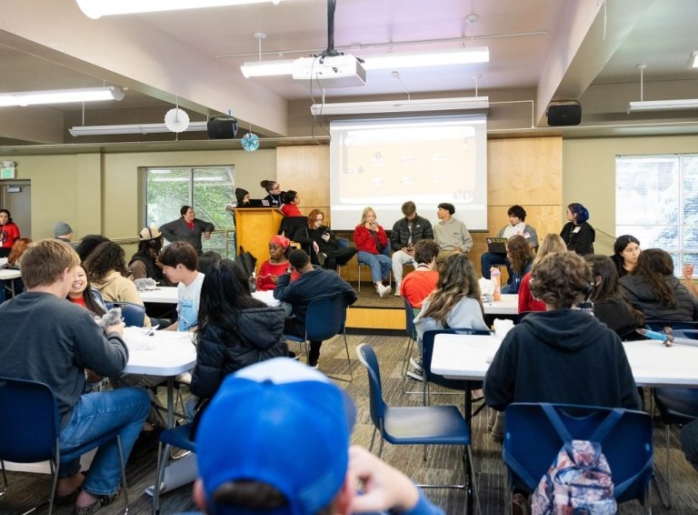 A large group of students sits while some sit in front of a screen at front of the room